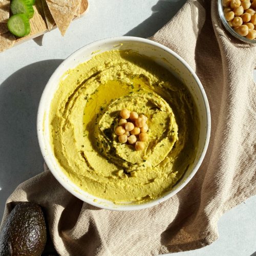 creamy avocado hummus in a small bowl topped with olive oil and extra chickpeas in the sunlight