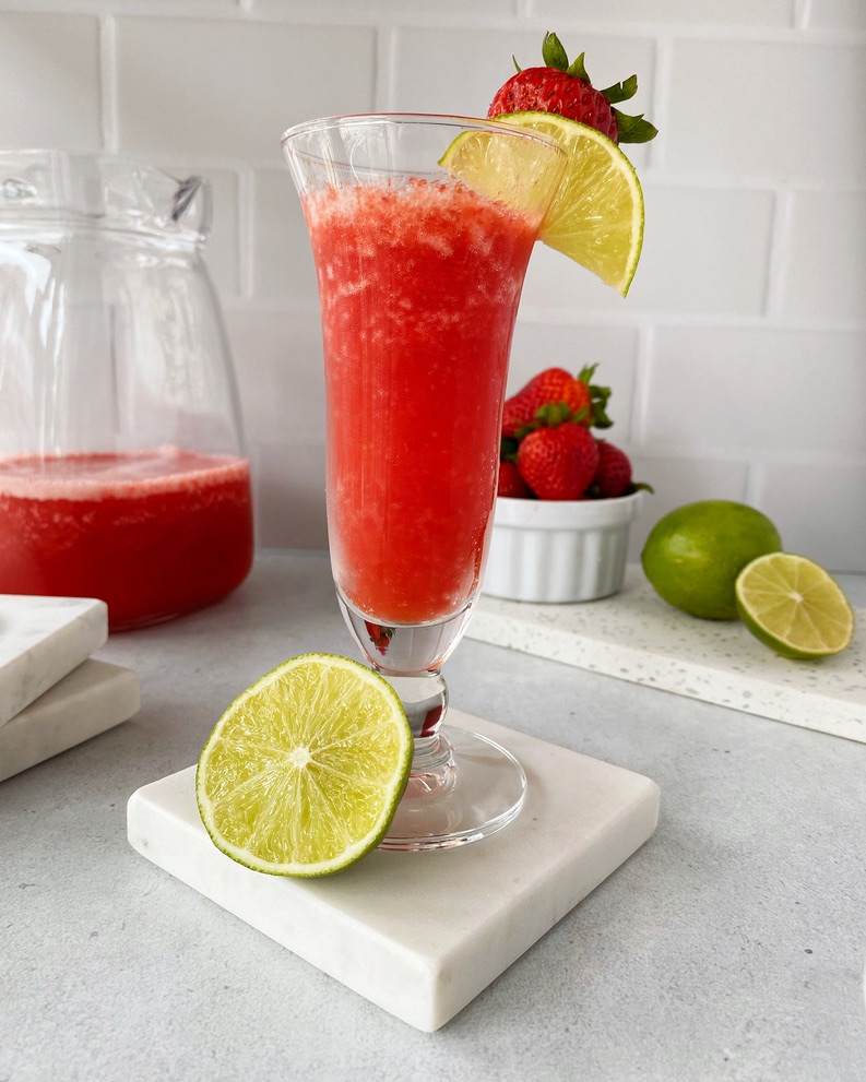 Strawberry daiquiri in a tall thin glass placed on a marble coaster with half a lime resting on the side of the glass