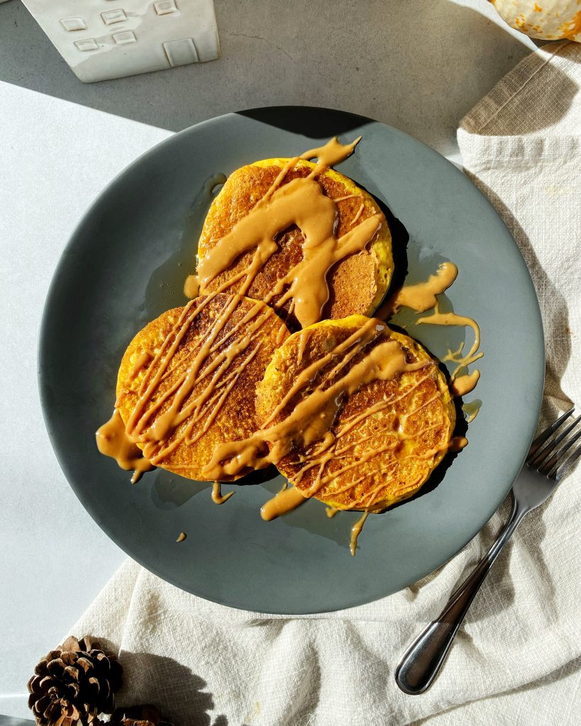 overhead shot of three pumpkin pancakes on a dark grey plate drizzled with peanut butter and maple syrup