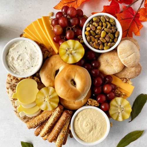 overhead shot of charcuterie board filled with vegan cheese, grapes, mini bagels, breadsticks, and pita bread